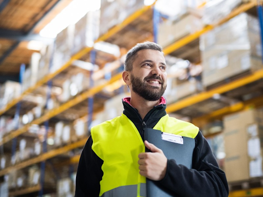 A man wearing a yellow safety vest is smiling and holding a clipboard inside a warehouse